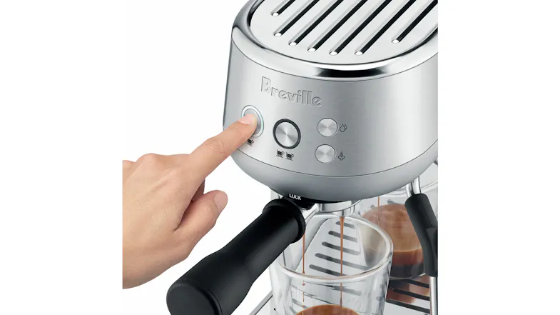 A hand pressing the brew button on the Breville Bambino as espresso flows into two clear glass cups A hand pressing the brew button on the Breville Bambino as espresso flows into two clear glass cups