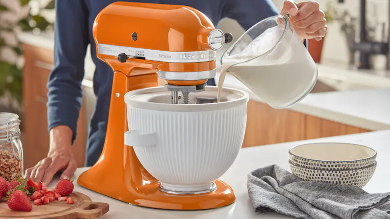 Pouring milk mixture into an orange KitchenAid stand mixer with the ice cream bowl attached Pouring milk mixture into an orange KitchenAid stand mixer with the ice cream bowl attached
