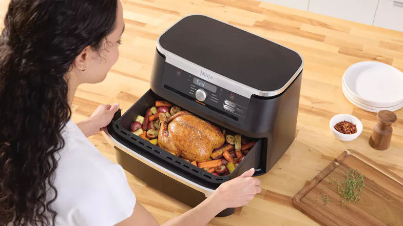 Person placing a roasted chicken and vegetables into an open Ninja air fryer on a kitchen counter Person placing a roasted chicken and vegetables into an open Ninja air fryer on a kitchen counter