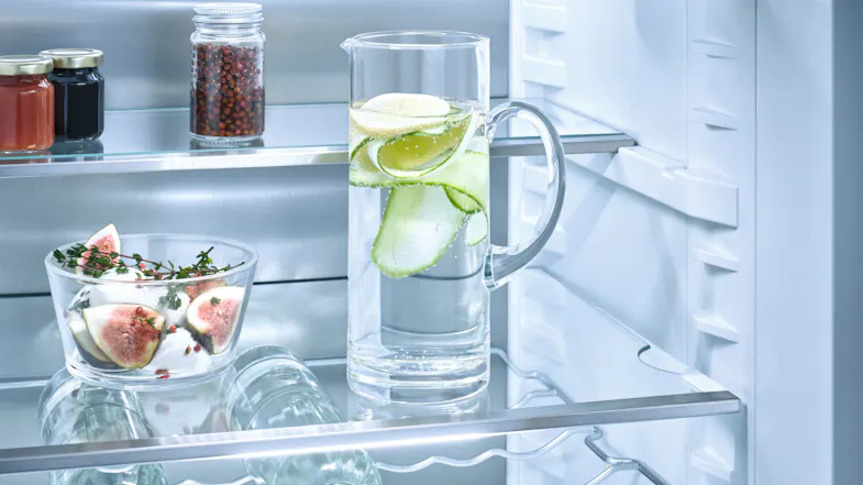 Glass shelves inside fridge holding a pitcher of water with cucumber slices, a bowl of figs, and jars Glass shelves inside fridge holding a pitcher of water with cucumber slices, a bowl of figs, and jars