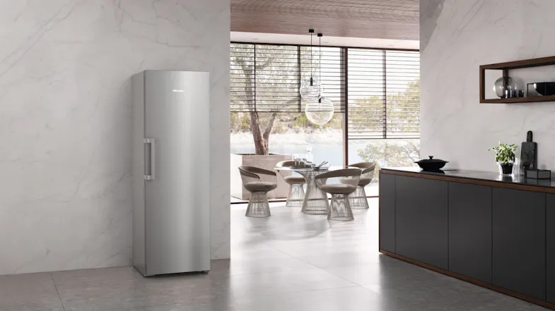 Miele 399L silver smart fridge standing in a modern kitchen with marble walls, dark cabinetry, and a dining area Miele 399L silver smart fridge standing in a modern kitchen with marble walls, dark cabinetry, and a dining area