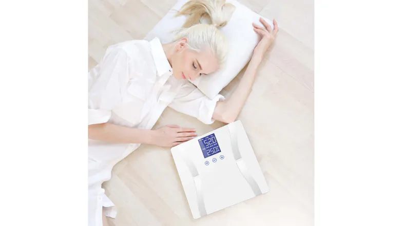 A person lying on the floor beside an electric weight scale displaying 52 kg and 25% body fat A person lying on the floor beside an electric weight scale displaying 52 kg and 25% body fat