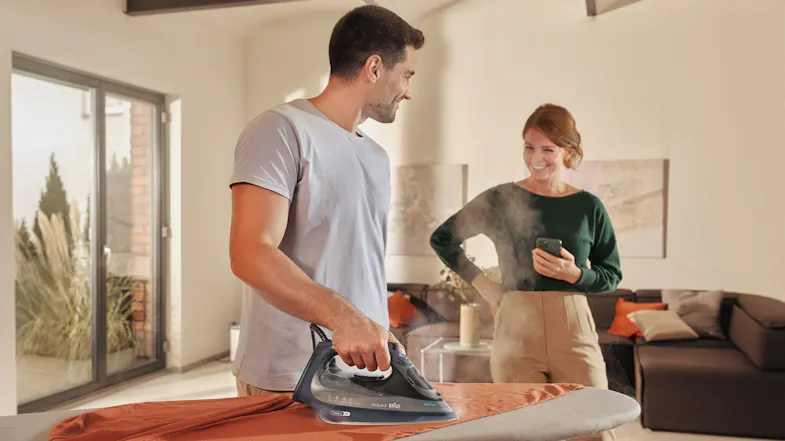 A person is ironing an orange shirt on an ironing board while another person stands nearby holding a smartphone in a living room A person is ironing an orange shirt on an ironing board while another person stands nearby holding a smartphone in a living room