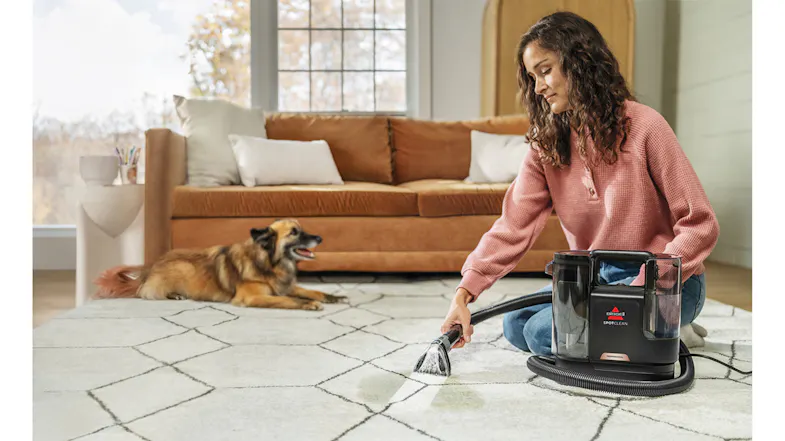 A person kneels on a rug using the SpotClean Professional tool to remove a stain while a dog watches nearby A person kneels on a rug using the SpotClean Professional tool to remove a stain while a dog watches nearby