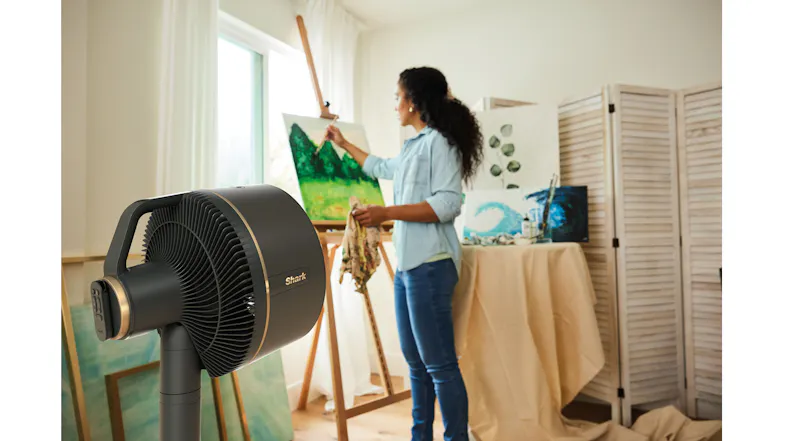 A pedestal fan is positioned in an art studio while a person paints on a canvas in the background A pedestal fan is positioned in an art studio while a person paints on a canvas in the background