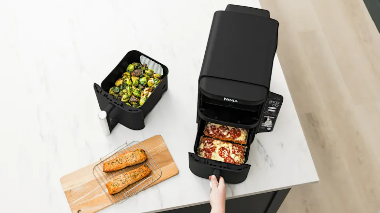 Hand pulling out an air fryer basket with cooked lasagna in one drawer and roasted vegetables in the other Hand pulling out an air fryer basket with cooked lasagna in one drawer and roasted vegetables in the other