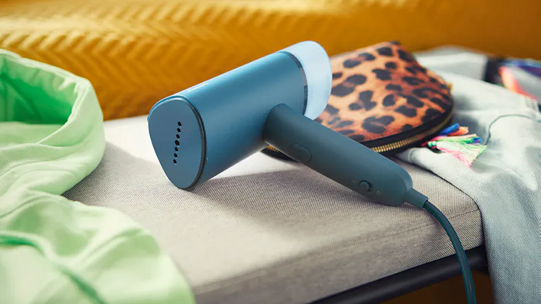 A handheld garment steamer in blue resting on a cushioned bench beside clothes and a leopard-print pouch A handheld garment steamer in blue resting on a cushioned bench beside clothes and a leopard-print pouch