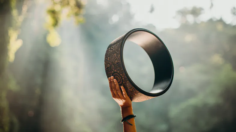 Hand holding a mandala yoga wheel outdoors with a sunlit forest backdrop for yoga and wellness practice Hand holding a mandala yoga wheel outdoors with a sunlit forest backdrop for yoga and wellness practice