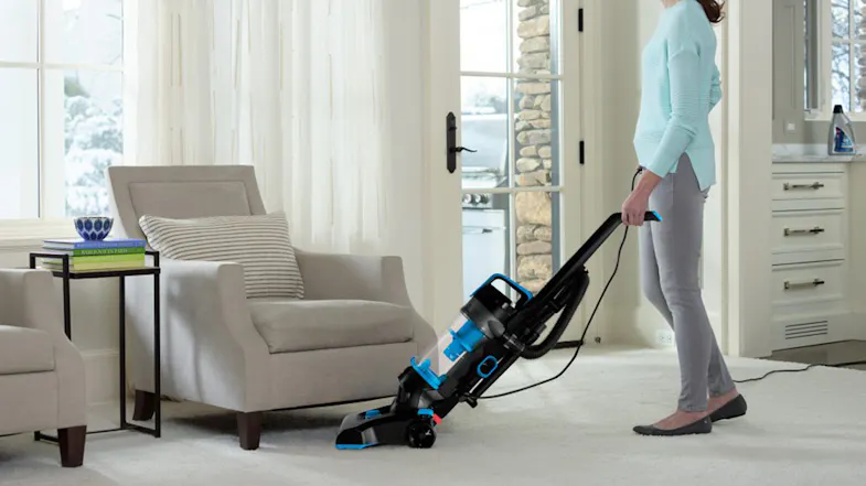 A person reclining with an upright vacuum to clean under a living room chair A person reclining with an upright vacuum to clean under a living room chair