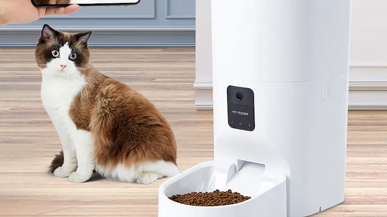 A cat is sitting beside a white automatic pet feeder with kibble in the bowl on a wood floor A cat is sitting beside a white automatic pet feeder with kibble in the bowl on a wood floor