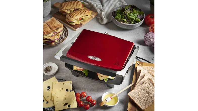 Red sandwich press cooking paninis, surrounded by bread, cheese, tomatoes, and salad Red sandwich press cooking paninis, surrounded by bread, cheese, tomatoes, and salad