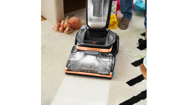 The Bissell Hydrosteam cleaner removes dirt and debris from a carpeted room scattered with toys The Bissell Hydrosteam cleaner removes dirt and debris from a carpeted room scattered with toys