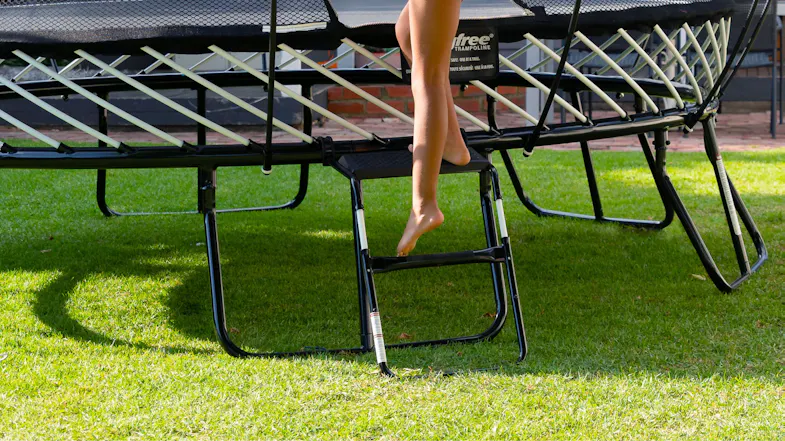 A child's foot stepping up on a small black metal ladder to climb into a Springfree trampoline on a lawn A child's foot stepping up on a small black metal ladder to climb into a Springfree trampoline on a lawn