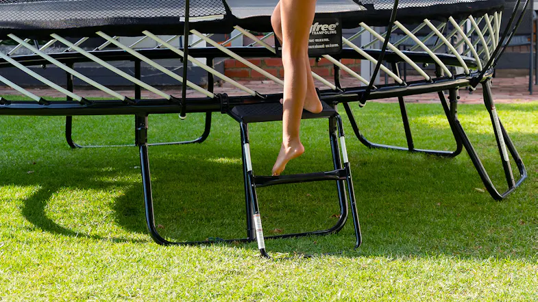 A child's foot stepping up on a small black metal ladder to climb into a Springfree trampoline on a lawn A child's foot stepping up on a small black metal ladder to climb into a Springfree trampoline on a lawn