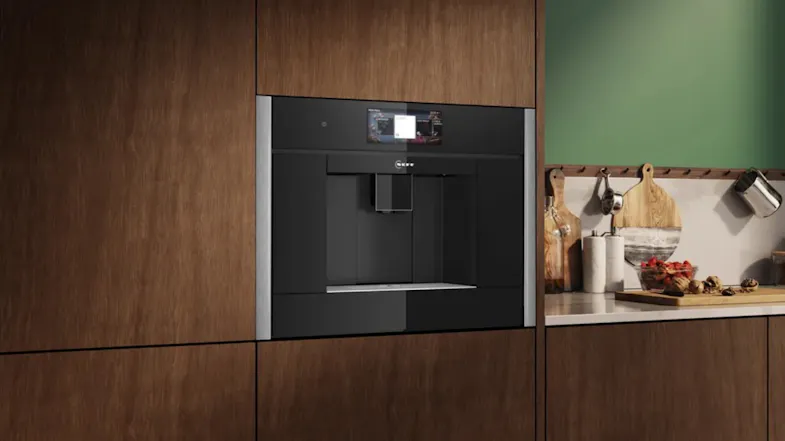 Built-in black automatic coffee machine installed in a modern wood-panelled kitchen beside a counter with cutting boards and utensils Built-in black automatic coffee machine installed in a modern wood-panelled kitchen beside a counter with cutting boards and utensils