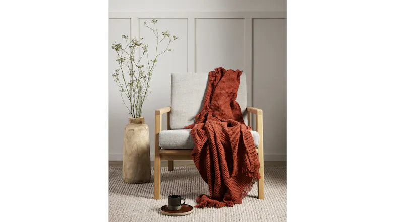 A brick-coloured throw draped over a light grey armchair beside a large vase with branches A brick-coloured throw draped over a light grey armchair beside a large vase with branches