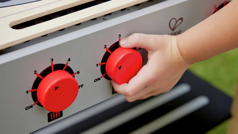 A close-up of a child's hand turning a red knob on a toy stove. A close-up of a child's hand turning a red knob on a toy stove.