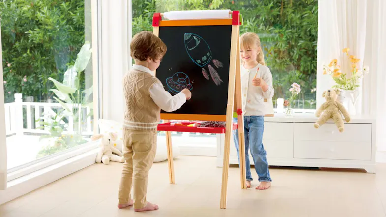 Two children drawing together on the blackboard side of the easel near a sunny window Two children drawing together on the blackboard side of the easel near a sunny window