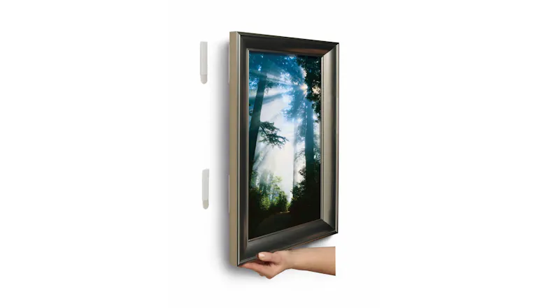 A hand hangs a framed forest photo on a wall using 3M Command Strips A hand hangs a framed forest photo on a wall using 3M Command Strips