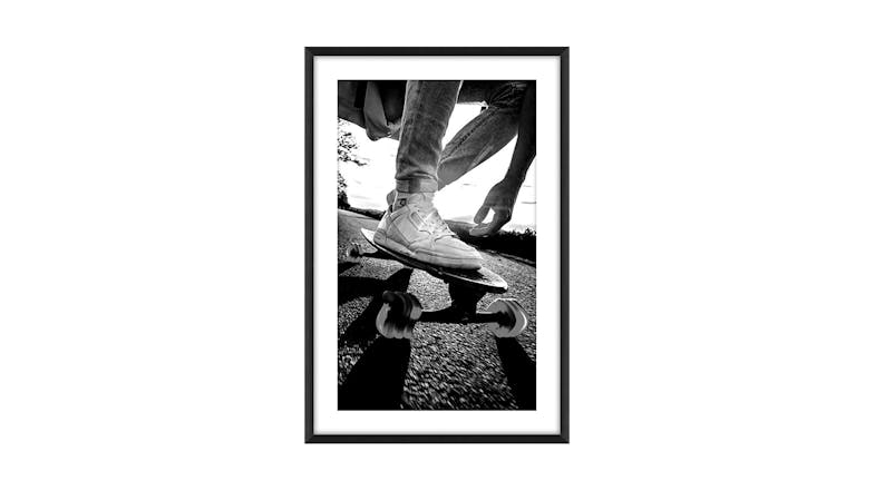 Low-angle black-and-white photo of skateboarder’s feet in sneakers riding on pavement, with shadows, trees, and sky in the background. Low-angle black-and-white photo of skateboarder’s feet in sneakers riding on pavement, with shadows, trees, and sky in the background.