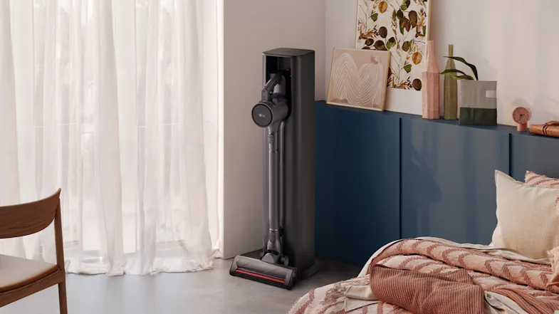 An LG handstick vacuum docked in an All-in-One Tower, charging in a bright, modern bedroom setting An LG handstick vacuum docked in an All-in-One Tower, charging in a bright, modern bedroom setting