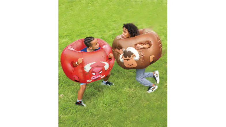 Two people wearing large inflatable bumper balls, one red with a bull face and one brown with a ram face, playing on grass Two people wearing large inflatable bumper balls, one red with a bull face and one brown with a ram face, playing on grass