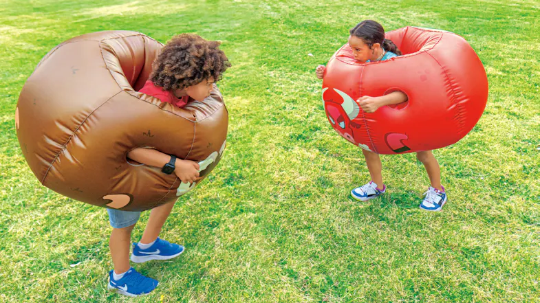 Two kids outdoors wearing large inflatable bumper balls, one brown with a ram face and one red with a bull face, ready to bump Two kids outdoors wearing large inflatable bumper balls, one brown with a ram face and one red with a bull face, ready to bump
