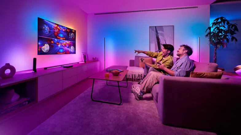 Two people seated on a sofa playing video games in a living room illuminated by Philips Hue lights Two people seated on a sofa playing video games in a living room illuminated by Philips Hue lights