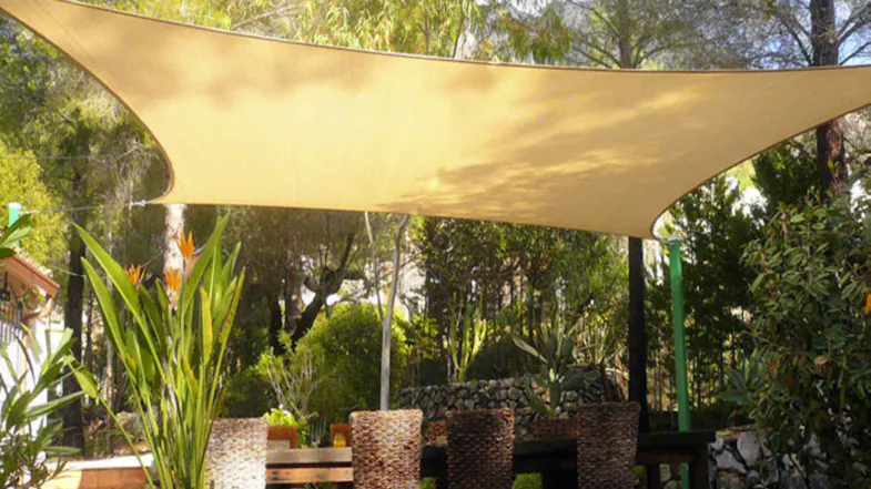 Outdoor dining setup with wicker chairs under a beige shade sail in a lush garden Outdoor dining setup with wicker chairs under a beige shade sail in a lush garden