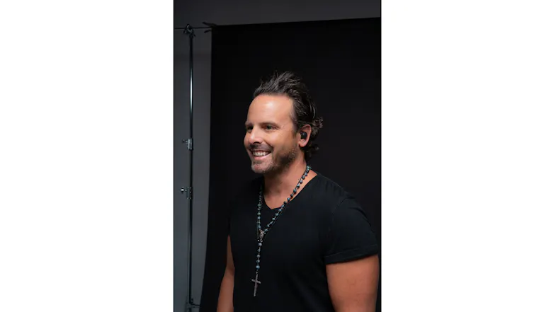 A person in a studio portrait wearing black earbuds and a cross necklace A person in a studio portrait wearing black earbuds and a cross necklace