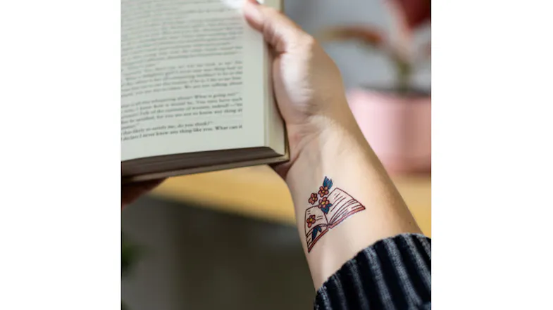 A person's forearm displays a colourful, custom-designed temporary book and flower tattoo while holding a book A person's forearm displays a colourful, custom-designed temporary book and flower tattoo while holding a book