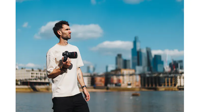 Photographer holding a Canon Mirrorless Camera by a river with a city skyline backdrop Photographer holding a Canon Mirrorless Camera by a river with a city skyline backdrop