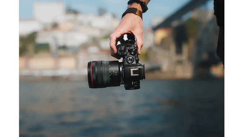 Hand holding a Canon EOS R6 Mark III with a mounted lens over a river Hand holding a Canon EOS R6 Mark III with a mounted lens over a river