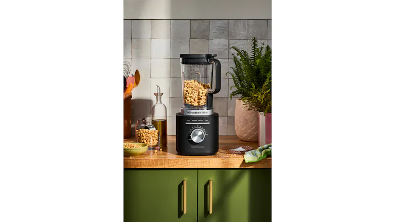 Blender with peanuts on a wooden counter, surrounded by kitchen items Blender with peanuts on a wooden counter, surrounded by kitchen items