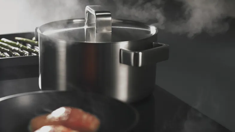 Cooktop in use with a stainless steel pot emitting steam and a tray of asparagus in the background Cooktop in use with a stainless steel pot emitting steam and a tray of asparagus in the background