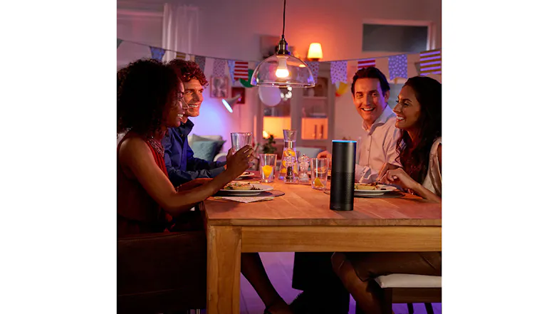 Warmly lit dining scene with four people, food, and a smart speaker on the table Warmly lit dining scene with four people, food, and a smart speaker on the table