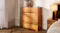 Full view of the rustic wooden four-drawer tallboy next to a bed with a lamp and decorative vases on top Full view of the rustic wooden four-drawer tallboy next to a bed with a lamp and decorative vases on top