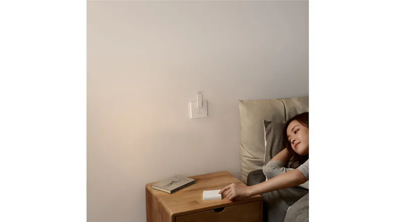 Aqara Hub M100 plugged into a wall socket near a wooden bedside table with books and a notepad Aqara Hub M100 plugged into a wall socket near a wooden bedside table with books and a notepad