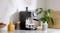 Close-up of a black espresso machine on a marble counter with two cups, a milk frother, juice, coffee beans, and a potted plant Close-up of a black espresso machine on a marble counter with two cups, a milk frother, juice, coffee beans, and a potted plant