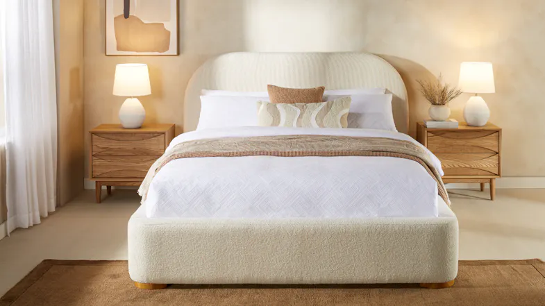 Front view of an upholstered bed with white textured bedding, decorative cushions, and a bedside lamp Front view of an upholstered bed with white textured bedding, decorative cushions, and a bedside lamp