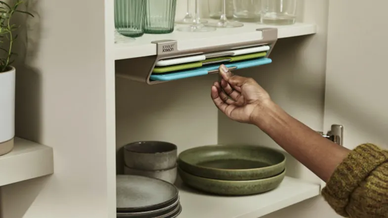 Hand pulling a colour-coded chopping board from Joseph Joseph under-shelf storage inside a kitchen cabinet Hand pulling a colour-coded chopping board from Joseph Joseph under-shelf storage inside a kitchen cabinet