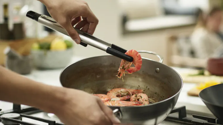 A hand is using stainless steel tongs to lift a cooked shrimp from a pan on a stovetop A hand is using stainless steel tongs to lift a cooked shrimp from a pan on a stovetop