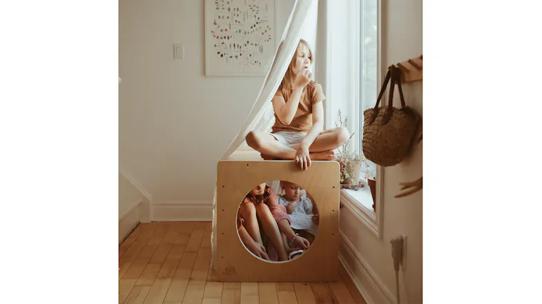 Three children playing in and on a wooden cube used as a cozy fort with a white sheet Three children playing in and on a wooden cube used as a cozy fort with a white sheet