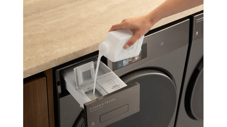 A hand pours liquid from a white bottle into a washing machine softener drawer A hand pours liquid from a white bottle into a washing machine softener drawer