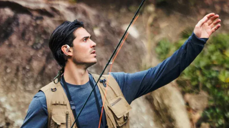 Man in a fishing vest wearing Shokz OpenFit Pro headphones while holding a fishing rod outdoors Man in a fishing vest wearing Shokz OpenFit Pro headphones while holding a fishing rod outdoors