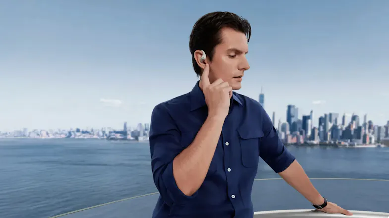 Man in a blue shirt touching his white Shokz OpenFit Pro earbud while standing overlooking a city harbor Man in a blue shirt touching his white Shokz OpenFit Pro earbud while standing overlooking a city harbor