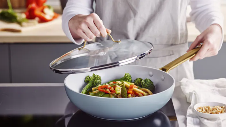 A person lifting the glass lid of a GreenPan wok to reveal a steaming vegetable stir-fry A person lifting the glass lid of a GreenPan wok to reveal a steaming vegetable stir-fry