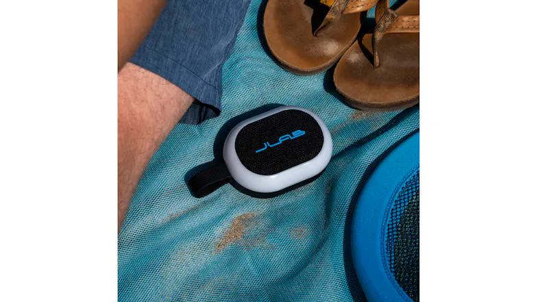 Overhead view of a JLab portable speaker on a blue beach blanket next to flip-flops Overhead view of a JLab portable speaker on a blue beach blanket next to flip-flops