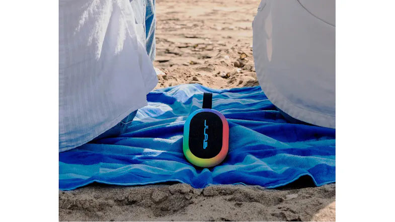 JLab speaker with a vibrant rainbow light ring sitting on a blue towel at a sandy beach JLab speaker with a vibrant rainbow light ring sitting on a blue towel at a sandy beach