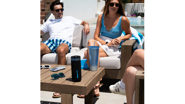 The JLab Go Party speaker sits on a wooden outdoor table while friends lounge by a pool The JLab Go Party speaker sits on a wooden outdoor table while friends lounge by a pool